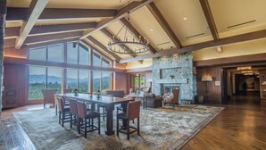 A spacious room with a large dining table, comfortable seating, a stone fireplace, and large windows offering a view of the mountains.