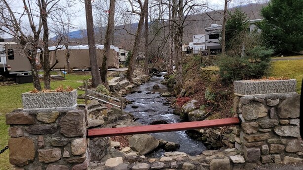 A stone bridge over a stream with a red railing, surrounded by trees and RVs in a campground setting. - Miss Daisy’s Retreat is a cozy getaway that will comfortably sleep four adults! (Gatlinburg)