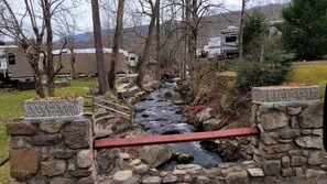 A stone bridge over a stream with a red railing, surrounded by trees and RVs in a campground setting.