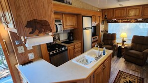 A compact RV kitchen with wooden cabinets, a stove, a sink, and a dining area with a chair and a table. - Miss Daisy’s Retreat is a cozy getaway that will comfortably sleep four adults! (Gatlinburg)