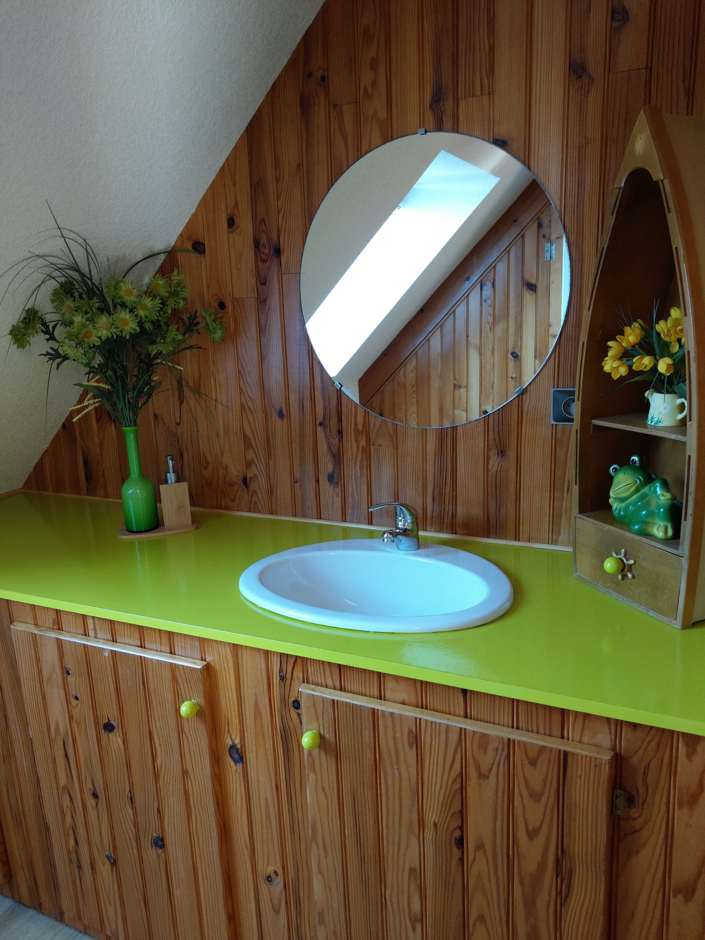 Un baño con tocador de madera, encimera verde, lavamanos blanco, espejo y un pequeño estante con una figurita de rana.