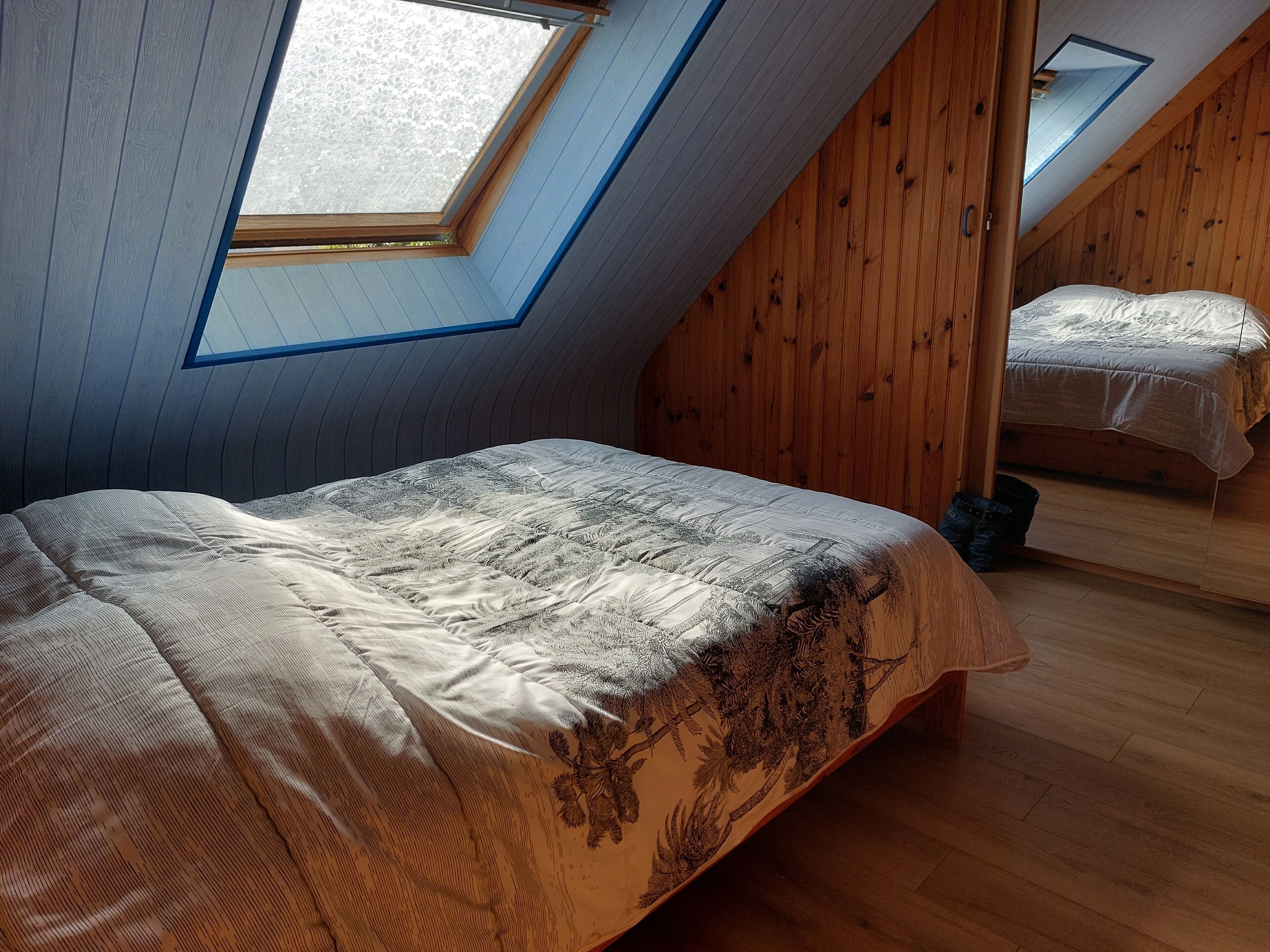 Une chambre avec un plafond en pente, des murs en bois, un lit recouvert d’une couvre-lit à motifs et une lucarne.