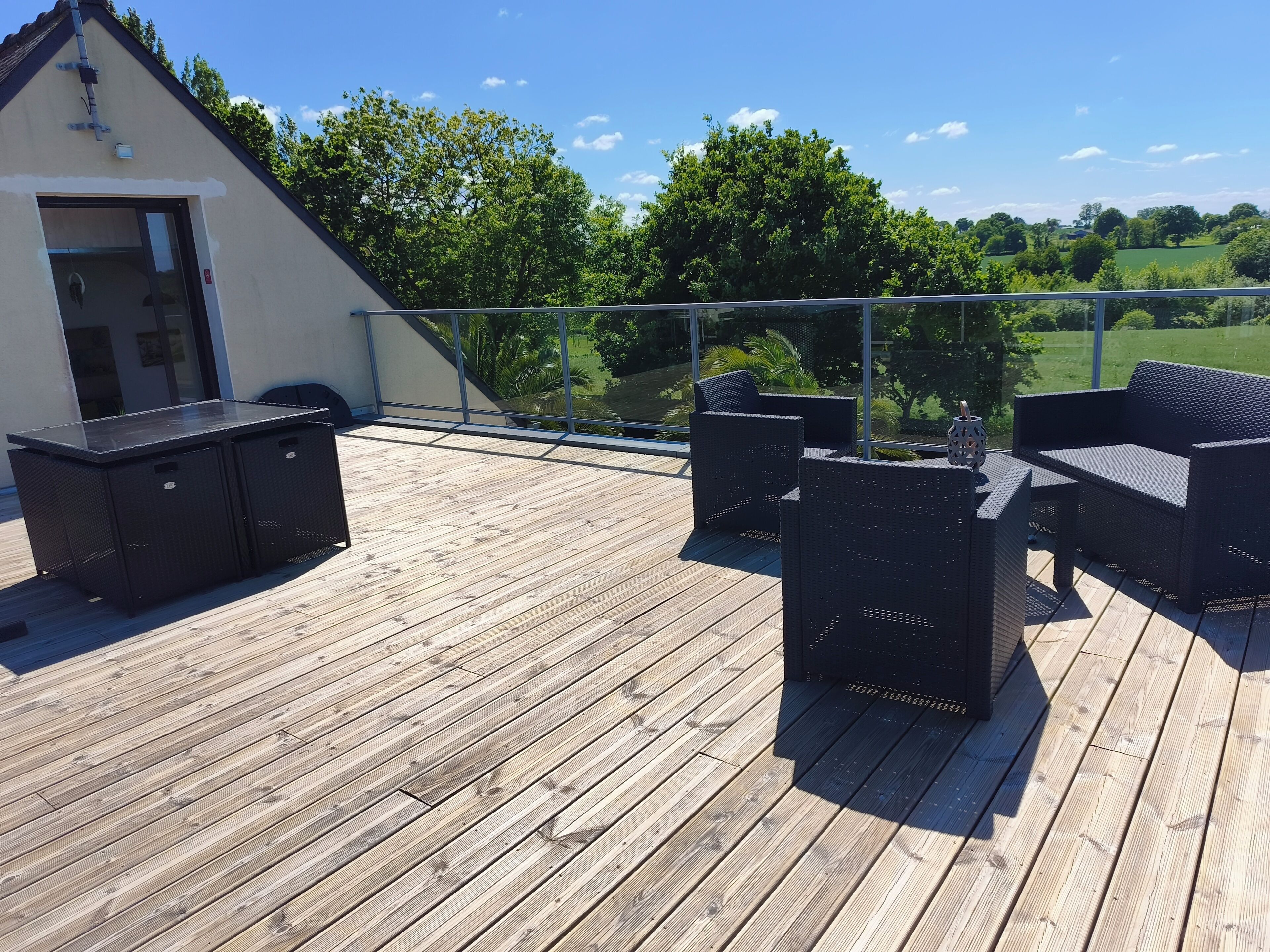 Une terrasse en bois, dotée de mobilier d’extérieur, d’une rambarde en verre et offrant une vue sur un paysage verdoyant.