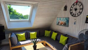 A modern living room with a glass coffee table, a sectional sofa with bright green cushions, a large wall clock, and a painting of candles. - Bel appartement avec terrasse à 5mn de SAINT-CAST (Matignon)