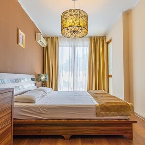 A bedroom with a wooden bed, a hanging light fixture, a bedside table, and a window with curtains. - Golden Line (Varna)