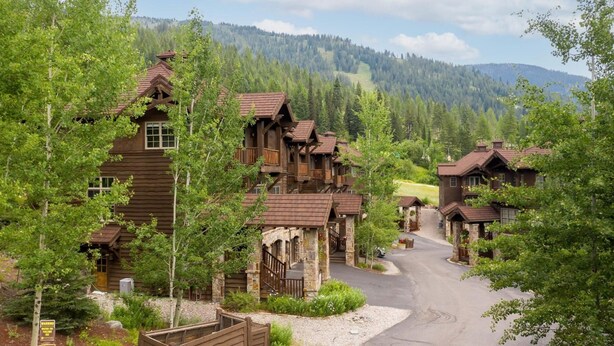 A rustic lodge with multiple wooden buildings, a stone entrance, and a road leading through a forested area with mountains in the background.