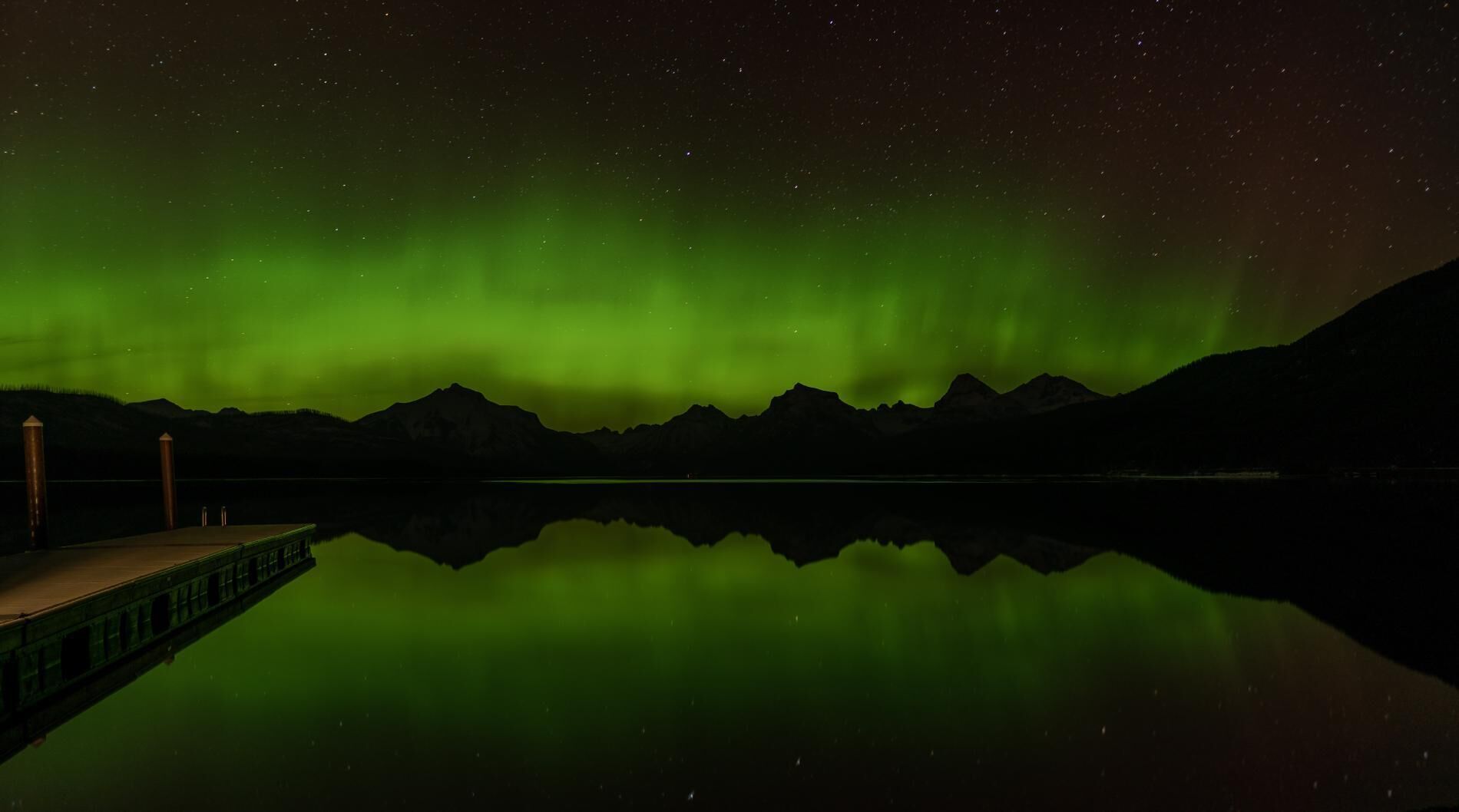 A night scene with the Northern Lights over a calm lake, reflecting the lights and mountains in the background.