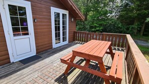Una terraza de madera con una mesa de picnic y bancos, una casa de madera con puertas blancas, y una valla de madera.