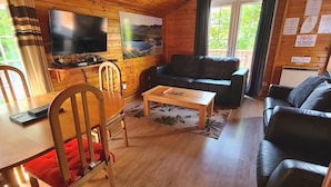 Un acogedor interior de cabaña con una mesa de comedor de madera, sillas, un sofá de cuero negro, un televisor montado en la pared y un ventanal con vistas a la vegetación.