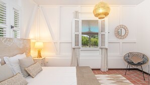 A bedroom with a bed, bedside table, lamp, window with shutters, and a chair.