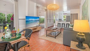 A spacious living area with a TV, wicker furniture, a dining table, and a kitchen in the background.