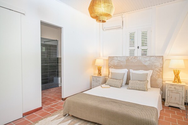 A bedroom with a large bed, two bedside tables with lamps, a wooden headboard with leaf patterns, and a door with shutters. - Bakwa (Le Vauclin)