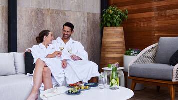 A couple in white robes sitting on a white sofa, a round table with drinks and snacks, and a large potted plant in a modern outdoor setting.