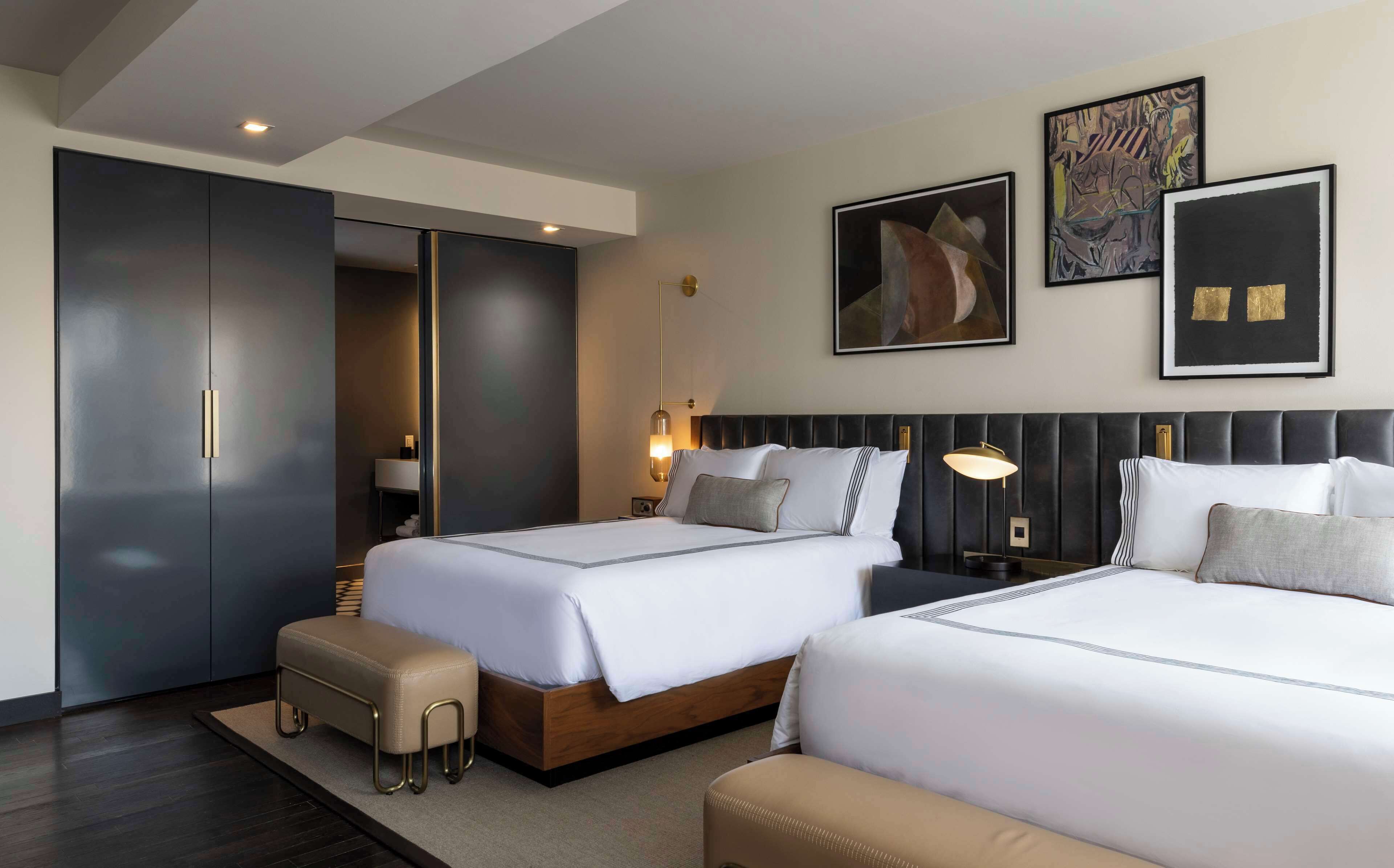 A hotel room with two beds, a wardrobe, a bedside table with a lamp, and artwork on the wall.
