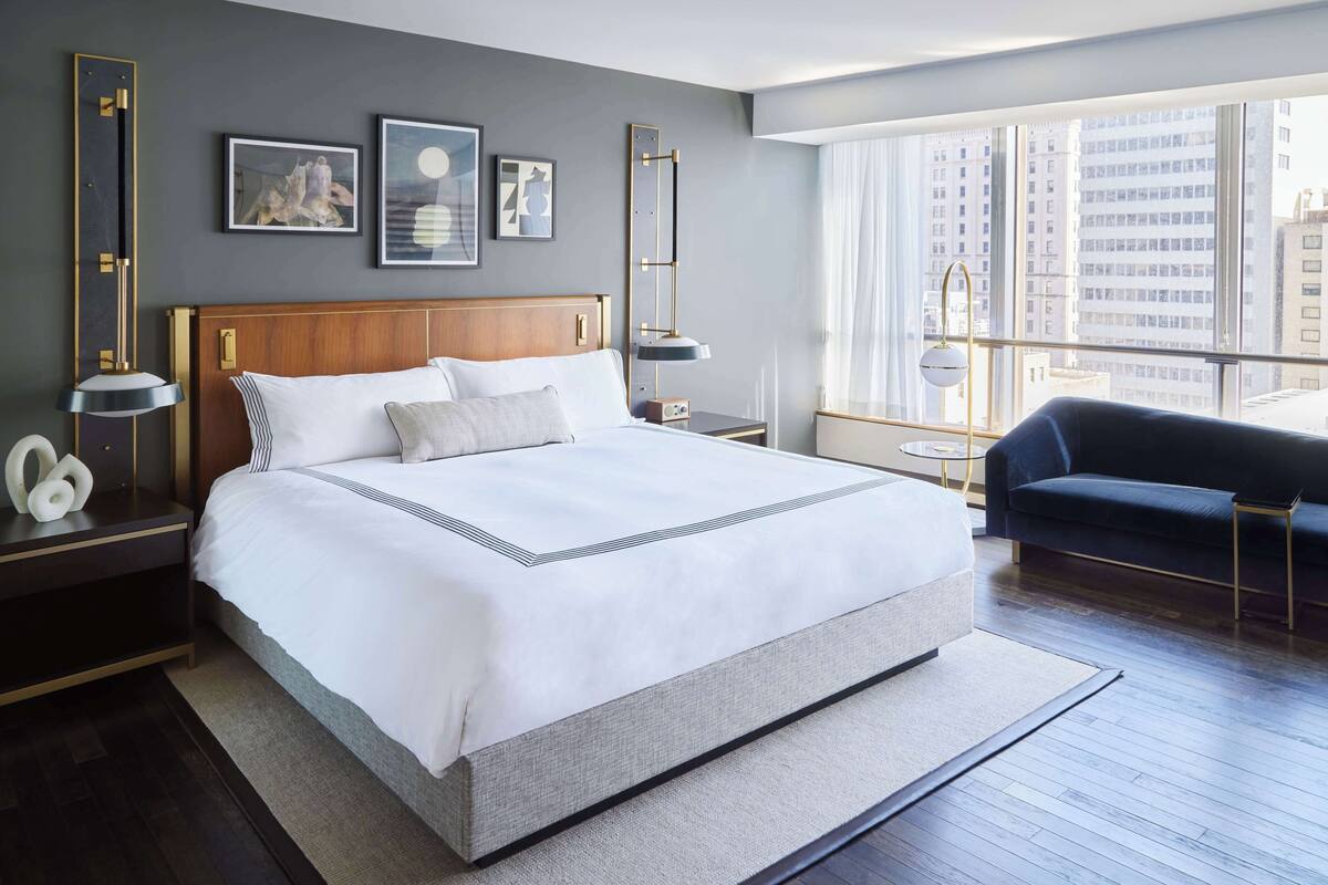 A modern hotel room with a large bed, a dark blue sofa, and a city view through large windows.