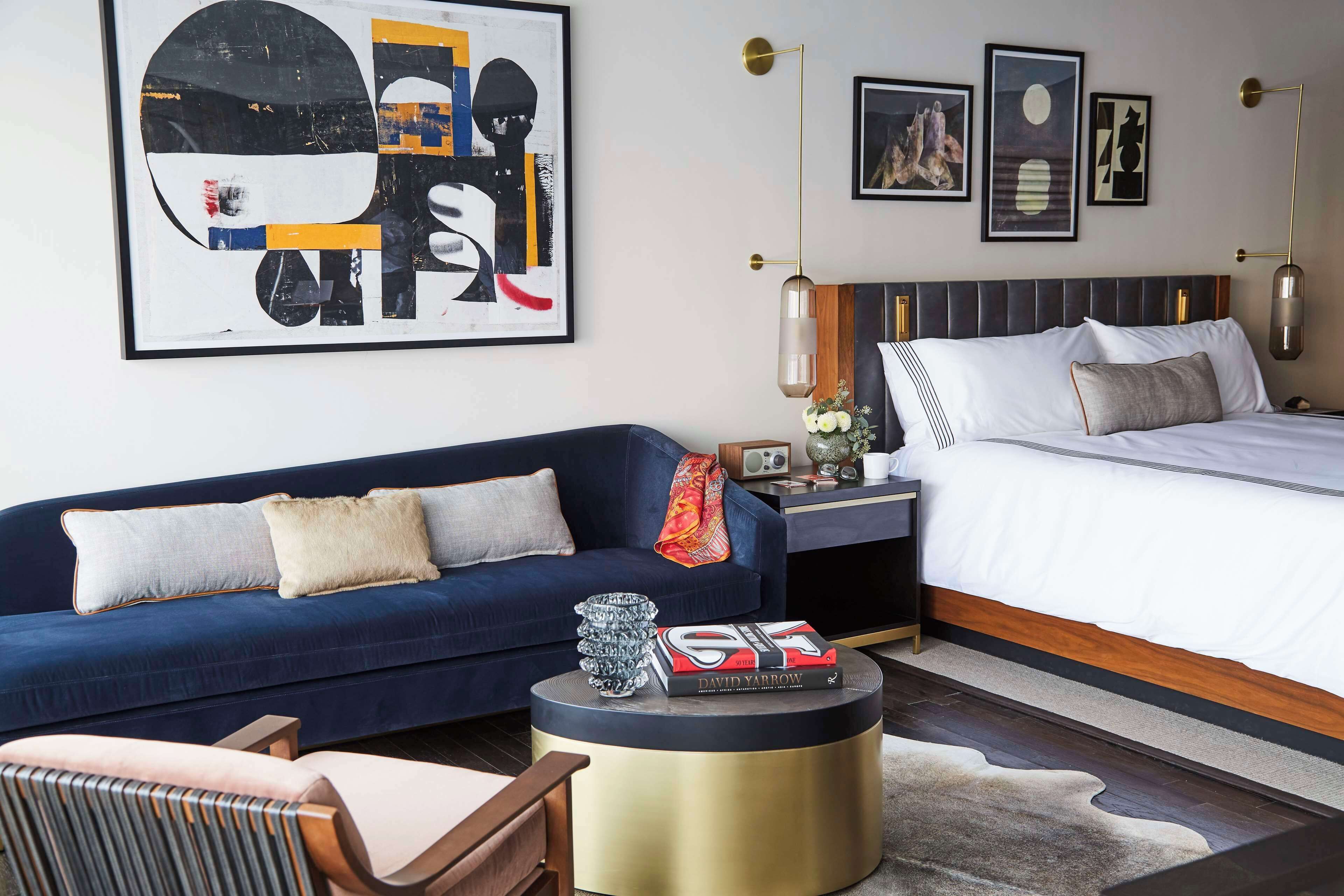 Una habitación de hotel moderna con un sofá azul, una mesa de centro redonda con libros, una cama con ropa de cama blanca y obras de arte en las paredes.