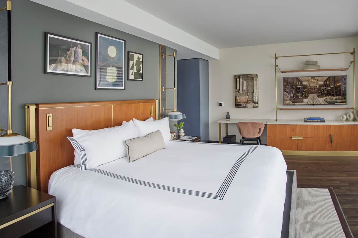 A hotel room with a large bed, a desk, and a chair. There are framed pictures on the wall and a large window with a view of the interior.