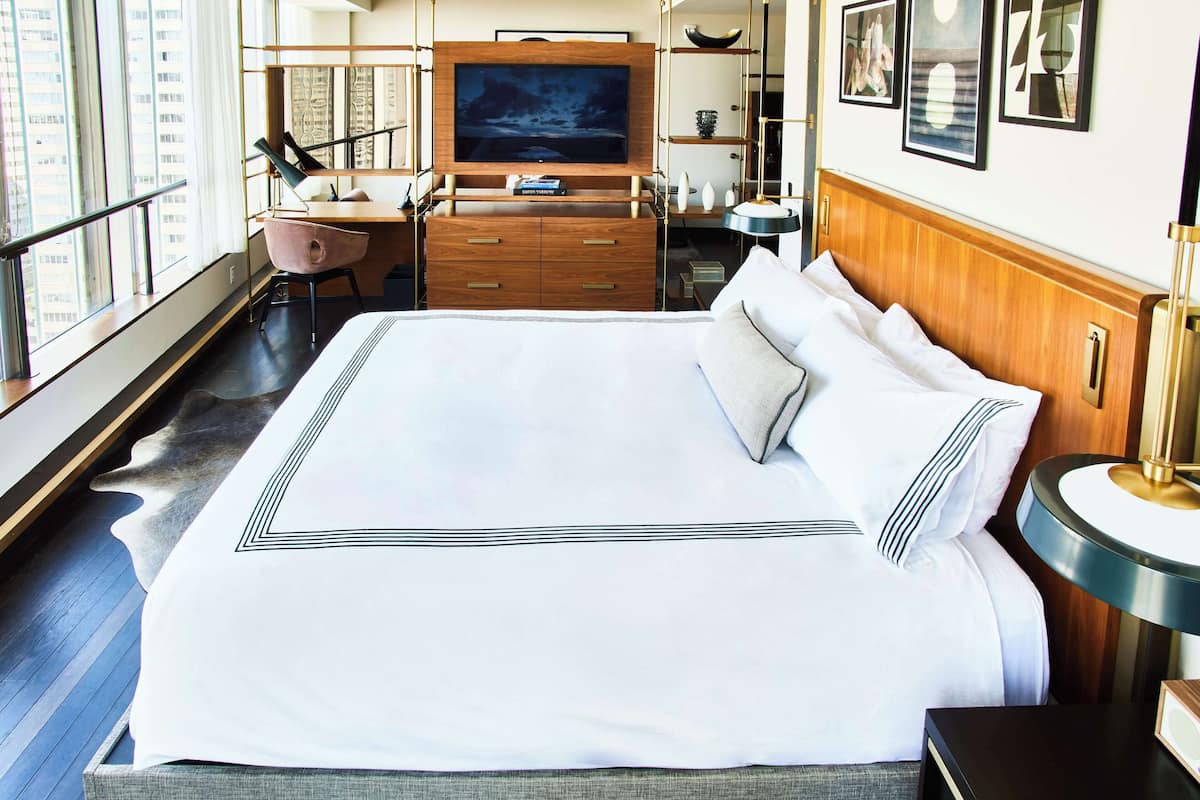 A hotel room with a large bed, a desk with a chair, a television, and a window with a view of buildings.
