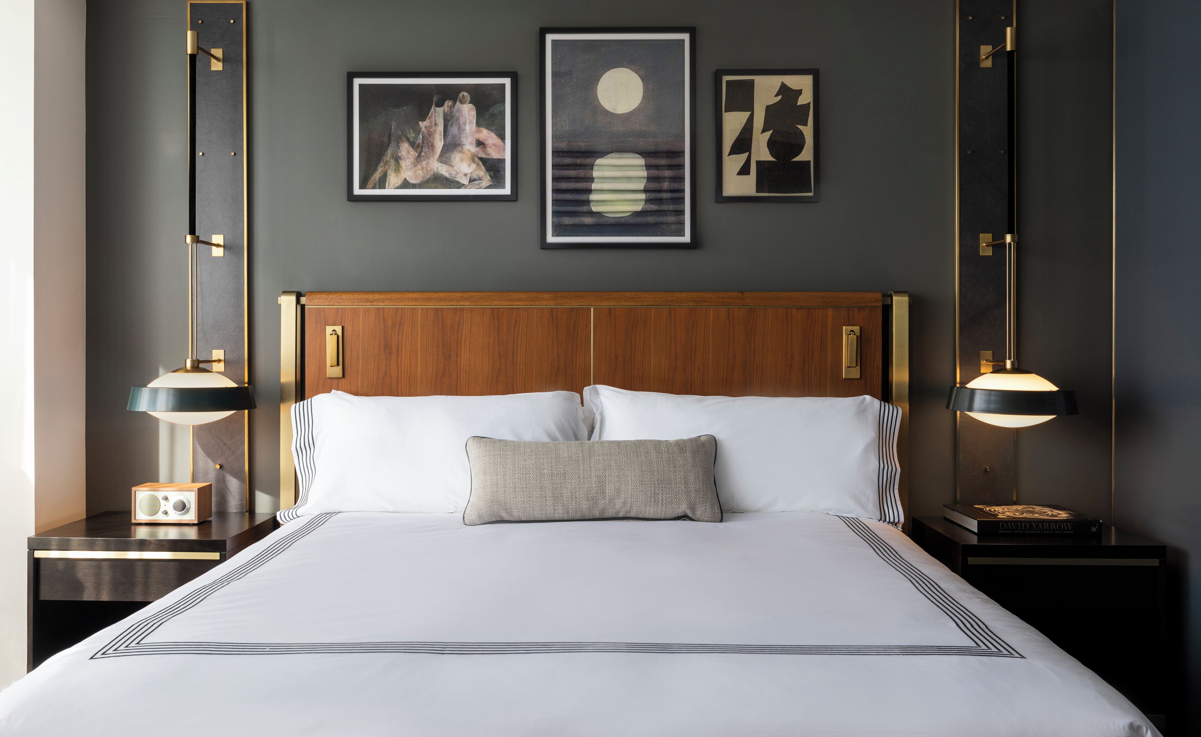 A neatly made bed with white linens and a wooden headboard, flanked by two bedside tables with lamps. Three framed artworks are hung on the wall above the bed.
