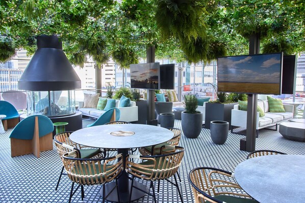 An outdoor seating area with round tables and wicker chairs, surrounded by potted plants and a large TV screen. - Thompson Dallas, by Hyatt (Dallas)