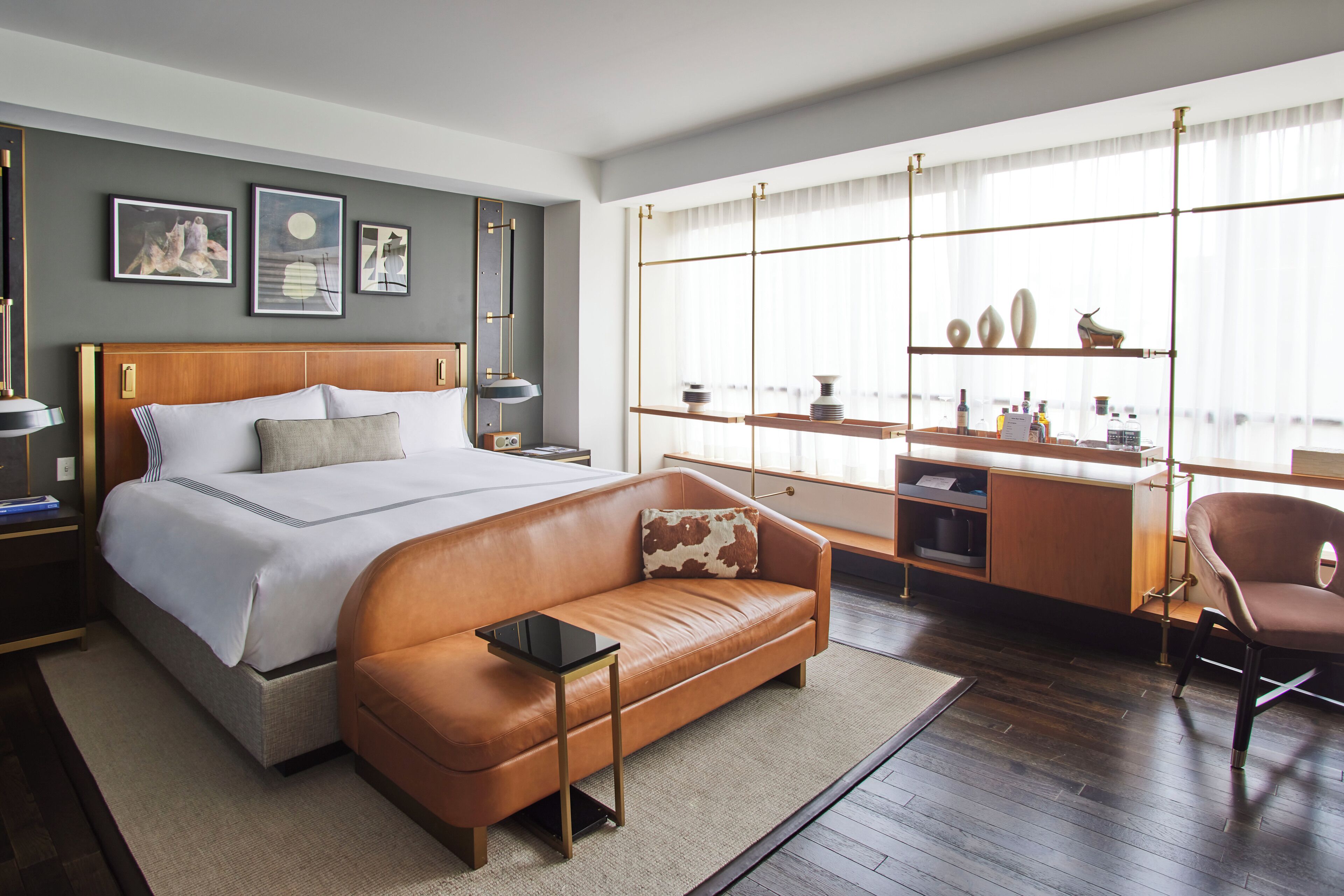 A modern hotel room with a large bed, a leather sofa, a desk with shelves, and artwork on the walls.
