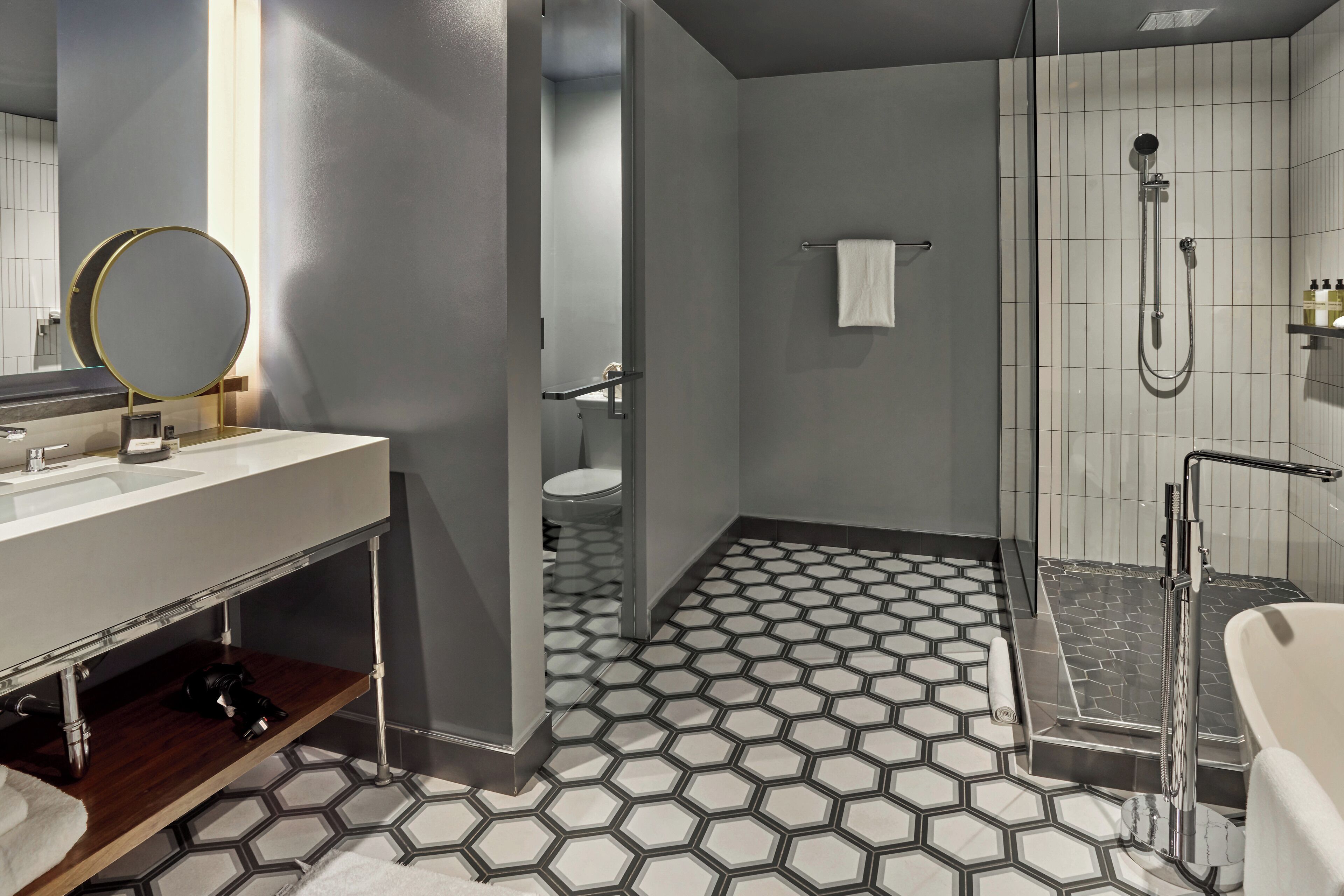 Une salle de bain moderne avec un sol carrelé à motifs hexagonaux, une douche en verre, une baignoire blanche et un meuble-lavabo agrémenté d’un miroir rond.