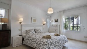A bedroom with a bed, bedside lamps, a dresser, a window with a view, and a hanging light fixture. - Villa Pinsapo (Benahavís)