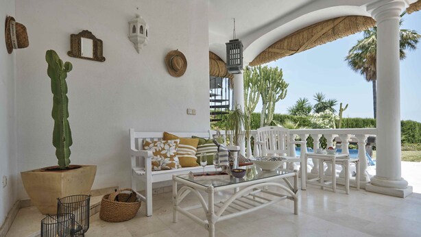 A white outdoor seating area with a glass table, white chairs, and a white bench. There is a large potted cactus, a wicker basket, and a hanging lantern. - Villa Pinsapo (Benahavís)