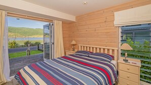 A bedroom with a large bed, a wooden headboard, a nightstand, and a window with blinds. - Opoutere Oasis - Opoutere Holiday Home (Opoutere)