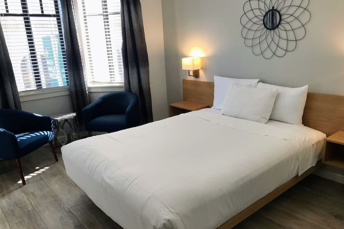 A hotel room with a large bed, a chair, a bedside table, a wall-mounted lamp, and a window with blinds.