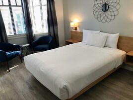A hotel room with a large bed, a chair, a bedside table, a wall-mounted lamp, and a window with blinds.