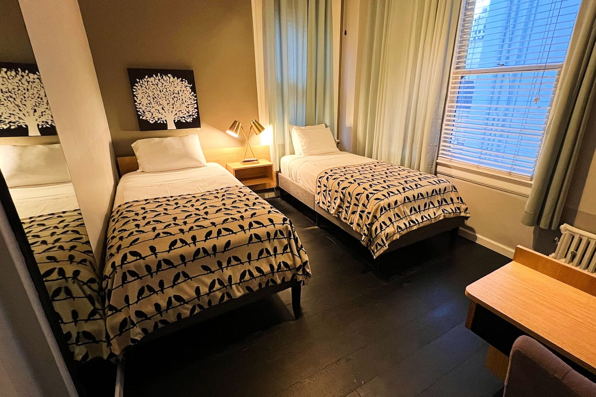A hotel room with two single beds, a desk, and a window with blinds.