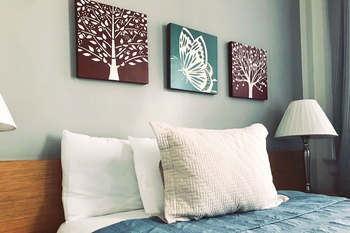 A hotel room with a bed, two pillows, a blue blanket, and three wall art pieces depicting trees and a butterfly.