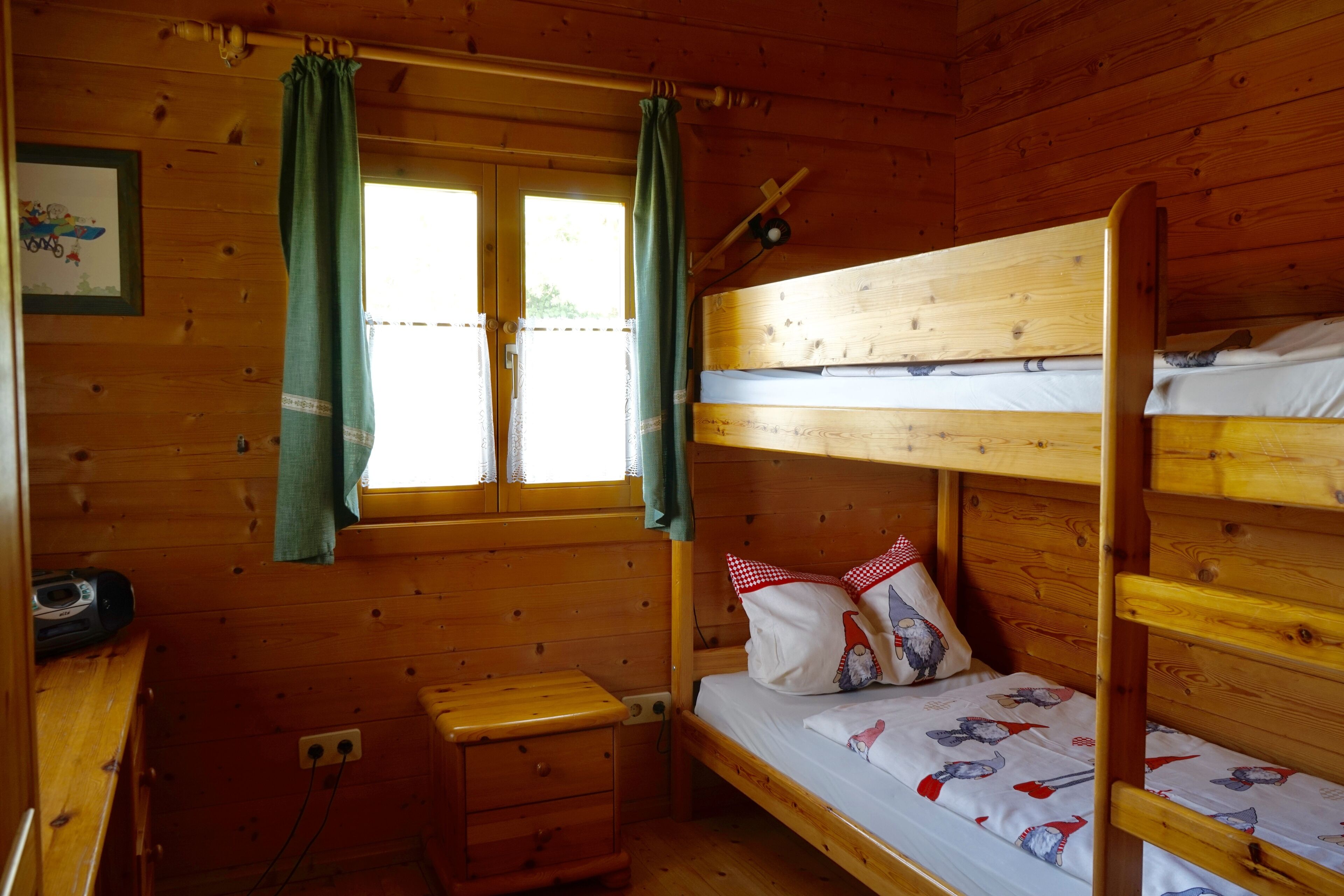A wooden cabin room with bunk beds, a small desk, and a window with curtains.