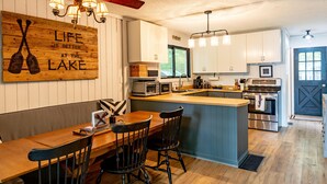 A kitchen with wooden cabinetry, a central island, and a dining area with chairs. - Adorable 3Bed, 2Bath Cottage and Garage Lounge! Grassy Lawns! Kayaks! (Rosseau)