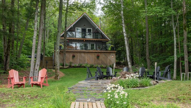 A cabin with a deck, surrounded by a forest. There are Adirondack chairs and a stone pathway leading to the cabin. - Adorable 3Bed, 2Bath Cottage and Garage Lounge! Grassy Lawns! Kayaks! (Rosseau)