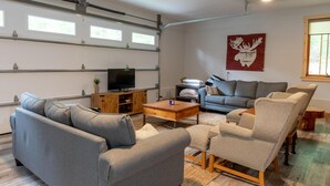 A modern living room with a grey sofa, a beige armchair, and a wooden coffee table. There is a flat-screen TV on a wooden stand, and a red moose artwork on the wall. - Adorable 3Bed, 2Bath Cottage and Garage Lounge! Grassy Lawns! Kayaks! (Rosseau)
