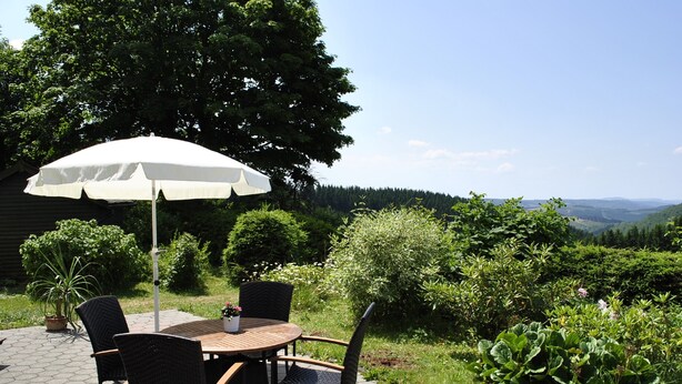 Um pátio externo com uma mesa redonda e cadeiras, um guarda-sol branco e vista para um vale arborizado.