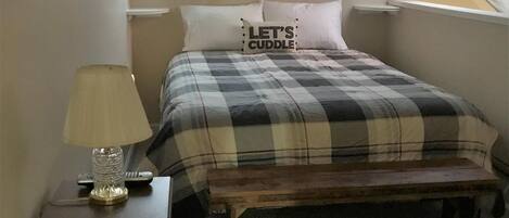 Um quarto com cama, mesa de cabeceira, luminária, um banco e janela.