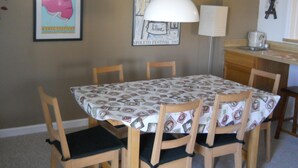 Un coin repas avec une table recouverte d’une nappe à motifs, entourée de chaises en bois dotées de coussins noirs.