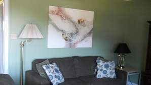 A living room with a grey sofa, two patterned cushions, a floor lamp, a table lamp, and a large abstract painting on the wall.