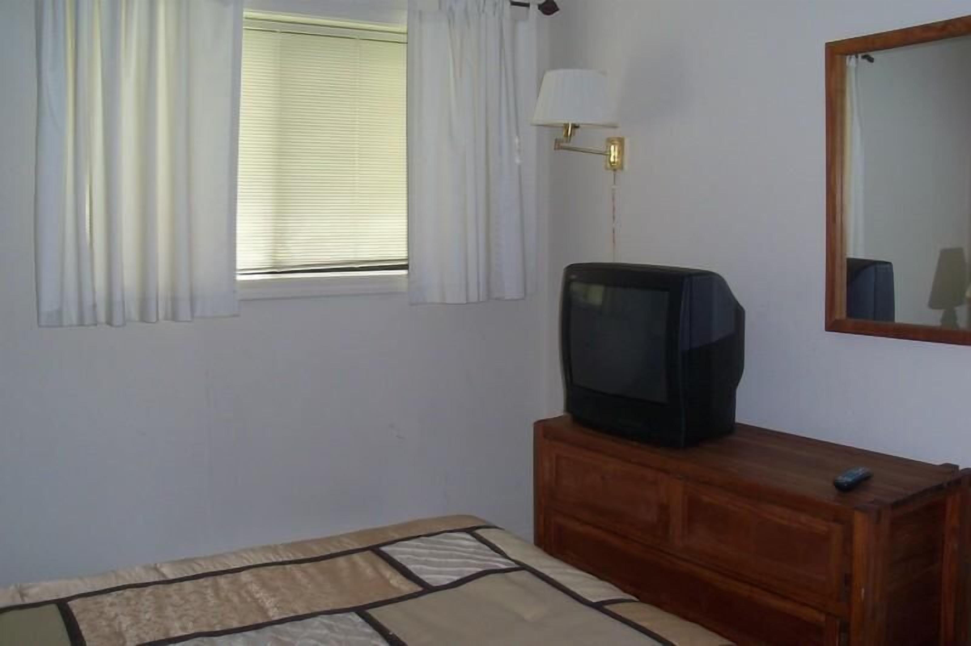 Quarto com cama, televisão sobre uma cômoda de madeira, janela com cortinas e luminária fixada na parede.