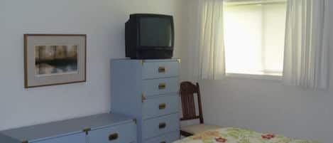 Un dormitorio con cama, cómoda, televisor encima de la cómoda, silla, ventana con cortinas e imagen enmarcada en la pared.
