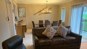 Ein Wohnzimmer mit einem braunen Ledersofa, zwei gleichfarbigen Sessel, einem Esstisch mit Stühlen und einer Deckenleuchte.