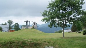Ländliche Landschaft mit einem kleinen Gebäude auf einem Hügel, umgeben von Bäumen und einigen Häusern im Hintergrund.