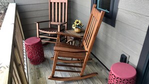A wooden rocking chair and a matching chair on a wooden deck with a small table and a vase of flowers.