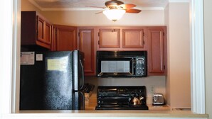Une cuisine de taille réduite, dotée de meubles en bois, d’un réfrigérateur noir, d’un four à micro-ondes, d’une cuisinière et d’un ventilateur de plafond.