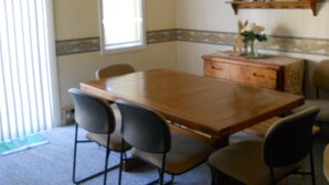 A dining area with a wooden table and four chairs. There is a window with blinds, a shelf with decorative items, and a patterned border on the wall.