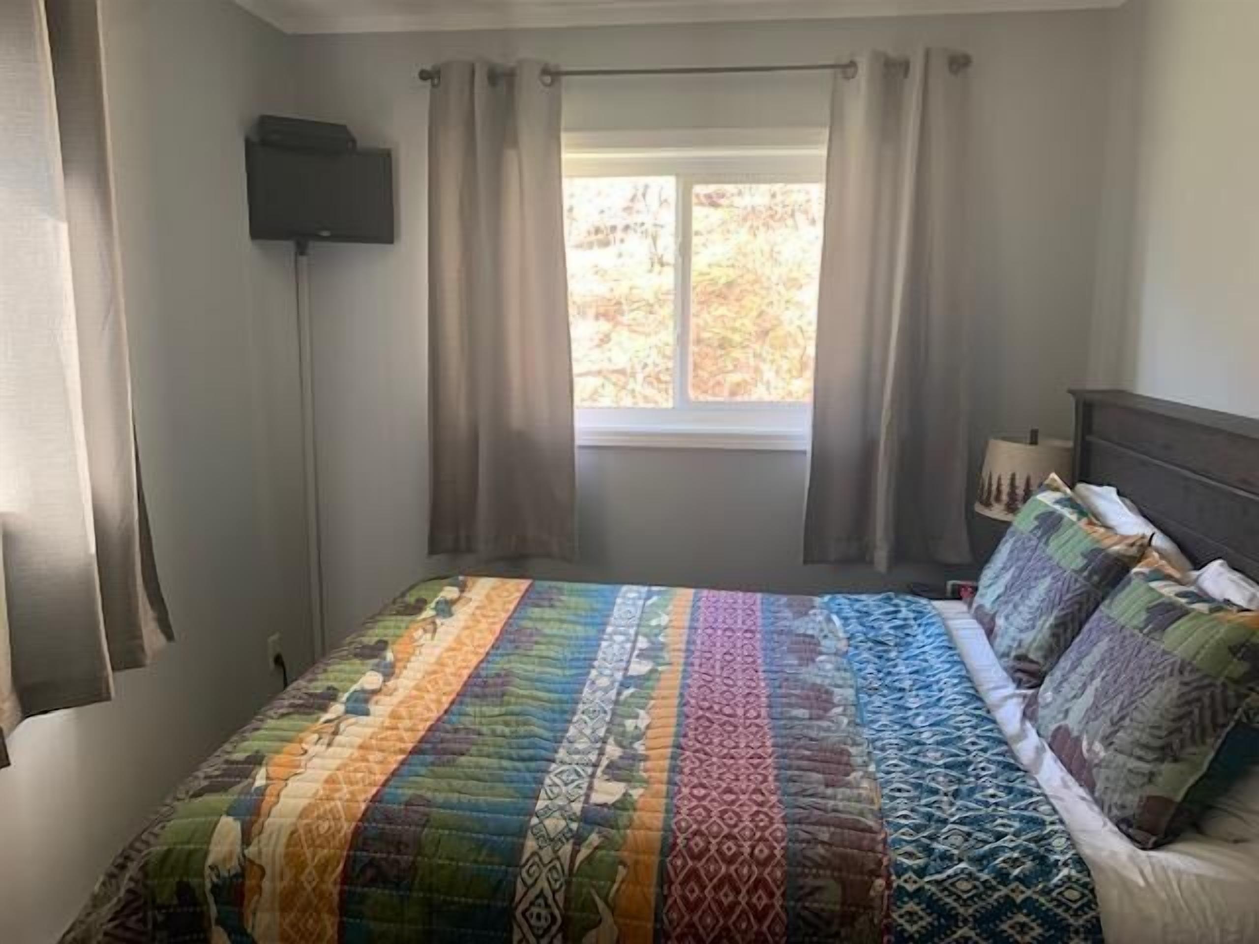 A bedroom with a large bed, a window with curtains, a bedside lamp, and a flat-screen TV mounted on the wall.