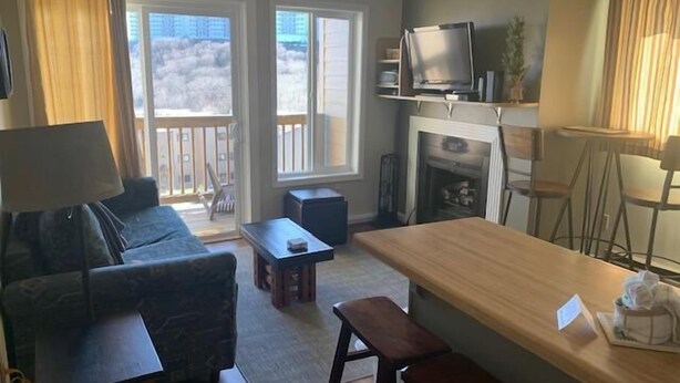 A living room with a sofa, a coffee table, a TV, and a fireplace. There are two windows with curtains, and a balcony is visible through one window. - Sugar Ski & Country Club Unit 125 Silver (Banner Elk)